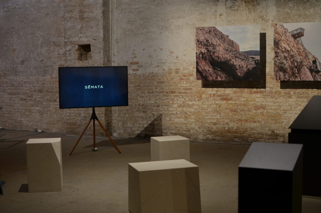 Exhibition with two photographs of the Akropolis in Athens, three stone seats facing an easel displaying the film “Sēmata (Signs)”, and a model of a ramp with slight vibrations.